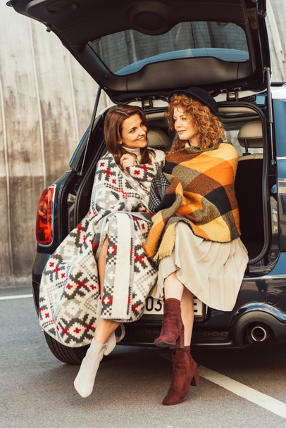 szczęśliwy dorosłe kobiety owinięte w koce, siedząc w bagażniku samochodu przy ulicy miejskiej - Zdjęcie, obraz