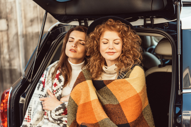 mujeres hermosas con los ojos cerrados envueltos en mantas sentadas en el maletero del coche en la calle
 - Foto, Imagen