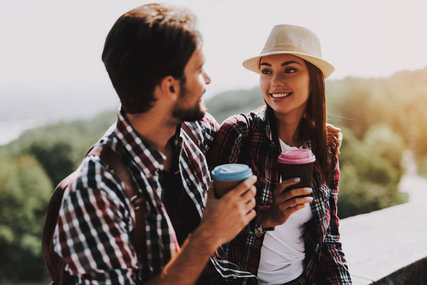 Happy Couple Sitting on Ledge and Drinking Coffee. Young Man and Woman with Backpacks Sitting Together on Concrete Ledge. Couple Enjoying Beautiful View of Nature. Traveling Concept - Foto, imagen