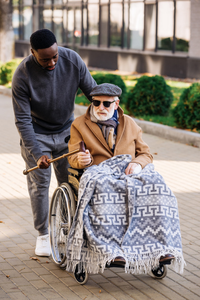 african american man giving walking stick to senior disabled man in wheelchair on street - Foto, immagini