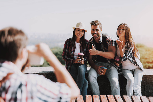 Young Man Taking Photo of Smiling People in Park. Group of Young Friends Sitting Together and posing for Group Photo. Young Travelers Enjoying Summer Sunny Day. Traveling and Friendship Concept - Foto, imagen