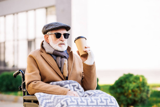 senior disabled man in wheelchair holding paper cup of coffee on street - Photo, Image