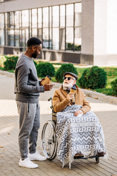 senior disabled man in wheelchair with plaid and african american man spending tome together on street with paper cups of coffee - Foto, immagini