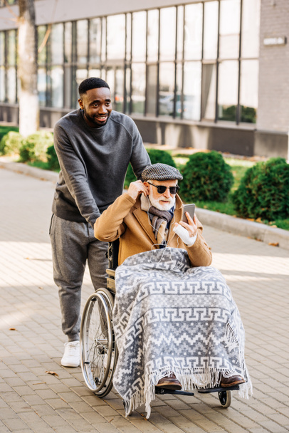 senior disabled man in wheelchair listening musing with smartphone and earphones with african american cuidador on street - Photo, Image
