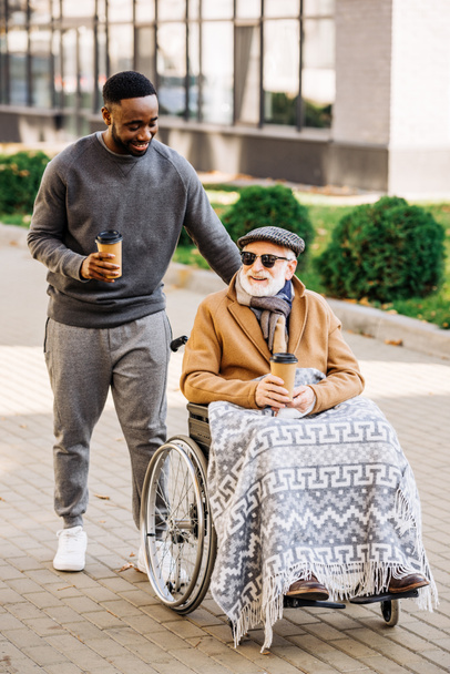 homme handicapé âgé souriant en fauteuil roulant avec plaid et homme afro-américain chevauchant par la rue avec des tasses en papier de café
 - Photo, image