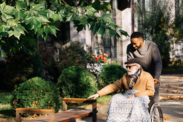 feliz idoso deficiente homem em cadeira de rodas e Africano americano cuidador passar tempo juntos na rua
 - Foto, Imagem