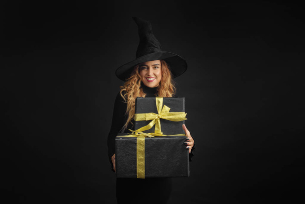 Black Friday. Halloween witch with a black hat on a black background holding gift boxes with juruse in her hands. Surprise and gift on sale - Foto, immagini