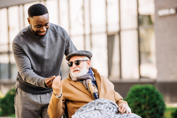 Afro-Amerikaanse man geven gezamenlijke senior gehandicapte man in de rolstoel wjhile ze samen tijd doorbrengen op straat - Foto, afbeelding