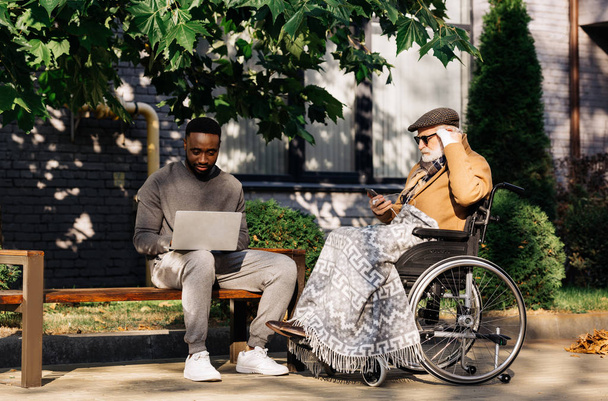 idoso deficiente homem em cadeira de rodas e homem afro-americano usando gadgets juntos na rua
 - Foto, Imagem