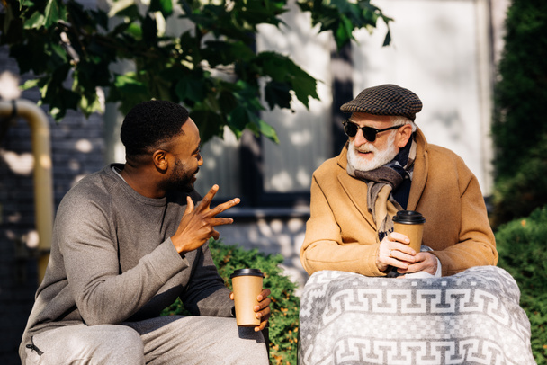 senior disabled man in wheelchair and african american man drinking coffee from paper cups and chatting on street - Foto, immagini