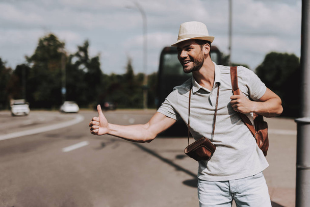 Young Tourist with Backpack Hitchhiking on Road. Casually dressed Handsome Man with Camera on Neck trying to Catch Car on Highway. Tourism and People Concept. Active Lifestyle. Summer Vacation - Foto, afbeelding