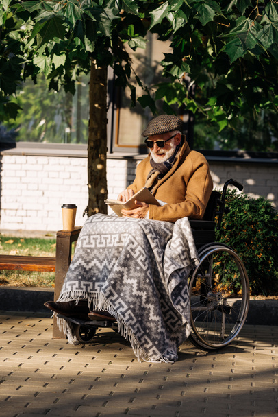 Senior uitgeschakeld man in rolstoel met papieren kopje koffie leesboek op straat - Foto, afbeelding