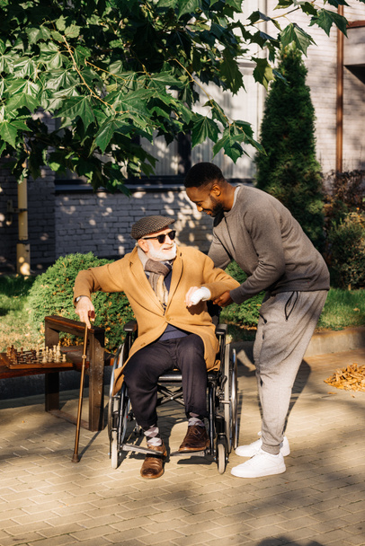 un homme afro-américain aidant un homme handicapé âgé à se lever de son fauteuil roulant dans la rue
 - Photo, image