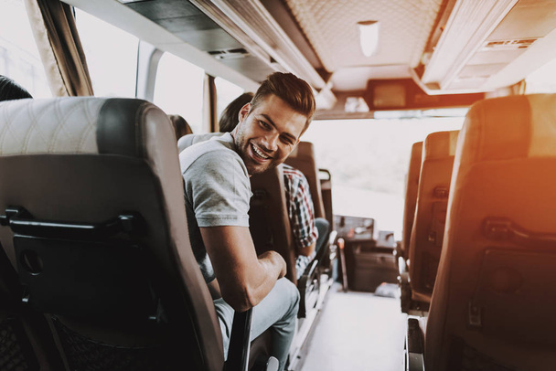 Hombre guapo divirtiéndose viajando en autobús turístico. Atractivo hombre sonriente sentado en el asiento del pasajero del autobús turístico y mirando hacia atrás. Concepto de Viajes y Turismo. Viajeros felices de viaje
 - Foto, imagen