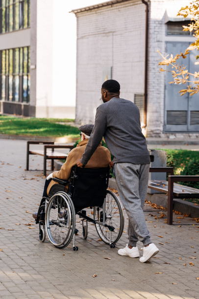 Achteraanzicht van senior gehandicapte man in de rolstoel en verpleegkundige rijden op straat - Foto, afbeelding