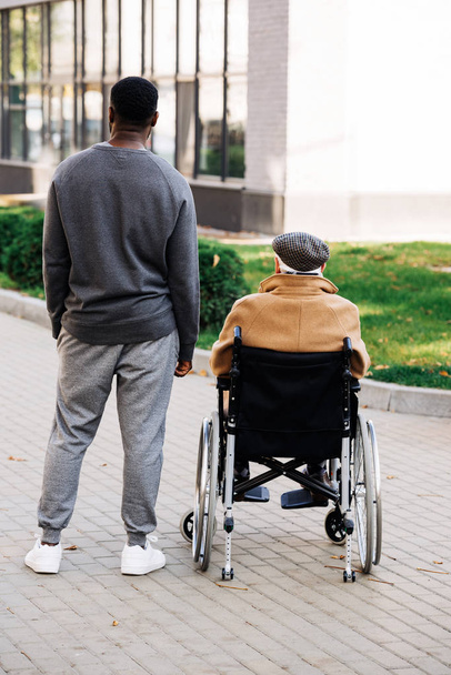 Rückansicht eines älteren behinderten Mannes im Rollstuhl und eines jungen afrikanisch-amerikanischen Cuidadors, die gemeinsam Zeit auf der Straße verbringen - Foto, Bild