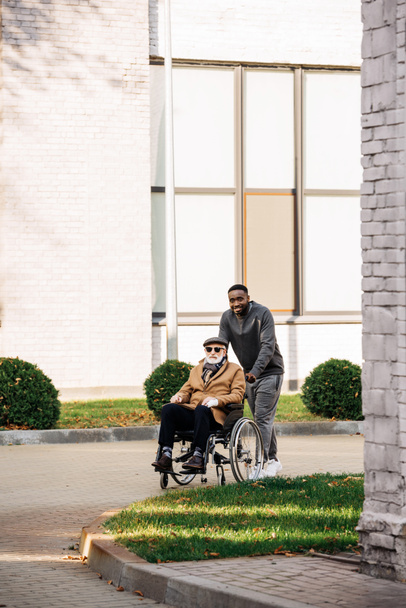 idoso deficiente homem em cadeira de rodas e afro-americano homem andando de rua juntos
 - Foto, Imagem
