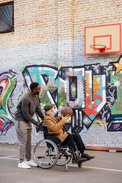 senior disabled man in wheelchair and happy african american man playing basketball together on street - Foto, Imagem