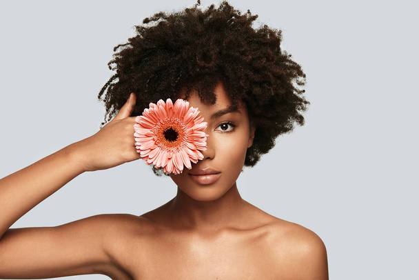 shirtless portrait of young African woman in studio looking at camera and covering eye with flower  - 写真・画像
