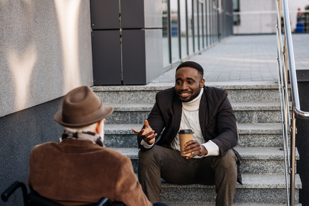 senior disabled man in wheelchair and african american man drinking coffee together and chatting on street - Photo, Image