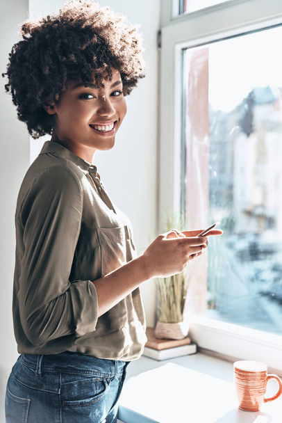 attractive young African woman using smart phone and smiling while standing near window indoors - Foto, Bild