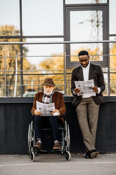 Senior gehandicapte man in rolstoel en knappe Afro-Amerikaanse man lezen kranten samen op straat - Foto, afbeelding