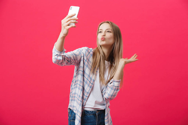 Γοητευτική νεαρή γυναίκα στο λευκό καπέλο ταξιδιού και Πάρτε selfie στην μπροστινή κάμερα smartphone που παρουσιάζουν απομονώνονται σε λάμψη ροζ φόντο. - Φωτογραφία, εικόνα