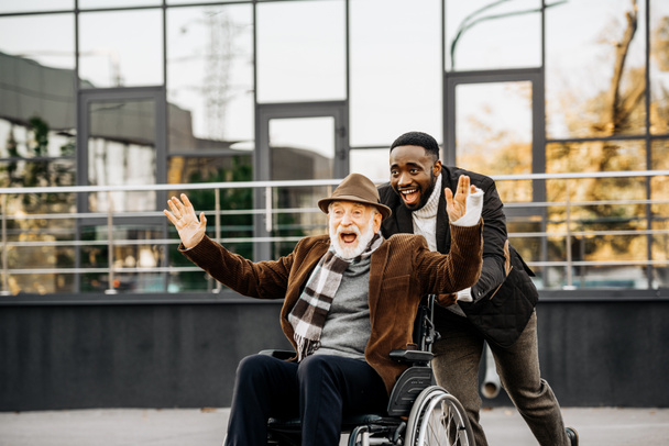 heureux homme handicapé senior en fauteuil roulant et homme afro-américain avoir du plaisir tout en chevauchant par la rue
 - Photo, image