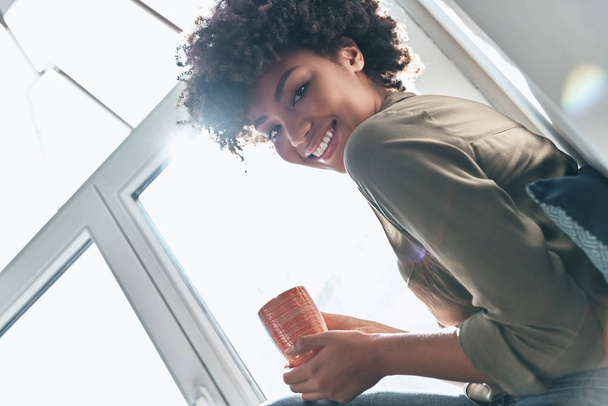 sonriente joven africana sosteniendo la taza de té en la ventana y mirando a la cámara
  - Foto, Imagen