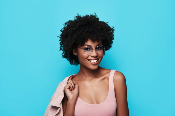 fashionable young African woman smiling and looking at camera while standing against blue background - Φωτογραφία, εικόνα