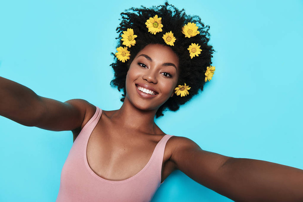 self portrait of attractive young African woman looking at camera and smiling while lying against blue background, yellow flowers in hair  - Foto, immagini