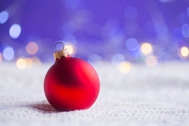 Bola de Navidad roja sobre tejido de punto blanco sobre fondo púrpura con bokeh cálido. Tarjeta de año nuevo. Decoración de Navidad
. - Foto, imagen