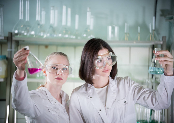 Chemical laboratory. Two young pretty woman holding different flasks with liquids in it. Looking at the flask - Photo, Image