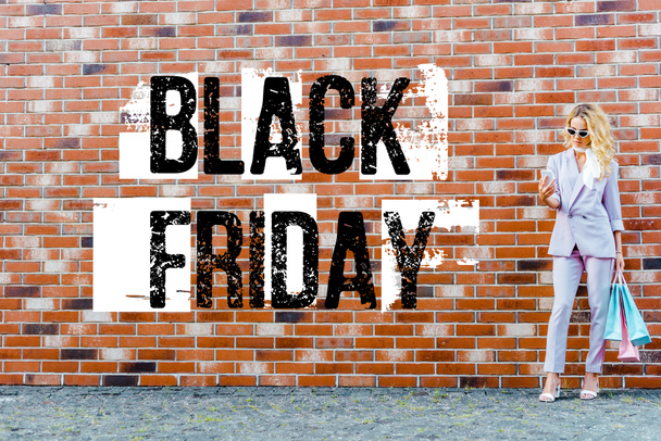 stylish young woman with shopping bags using smartphone while standing in front of brick wall, black friday banner - Photo, Image