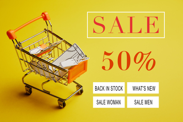 close up view of little shopping trolley with paper clothes on yellow backdrop, sale banner concept - Zdjęcie, obraz