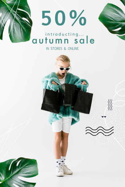 shocked kid in stylish fur coat looking into black shopping bags isolated on white, autumn sale banner concept - Photo, Image