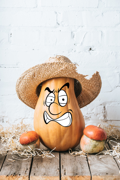 close up view of ripe pumpkins with drawn angry facial expression and straw hat on wooden surface and white brick wall backdrop - Fotoğraf, Görsel