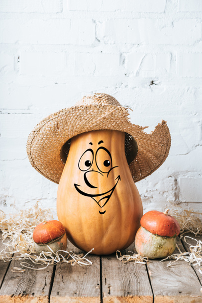 close up view of ripe pumpkins with drawn smiling facial expression and straw hat on wooden surface and white brick wall backdrop - Foto, Bild