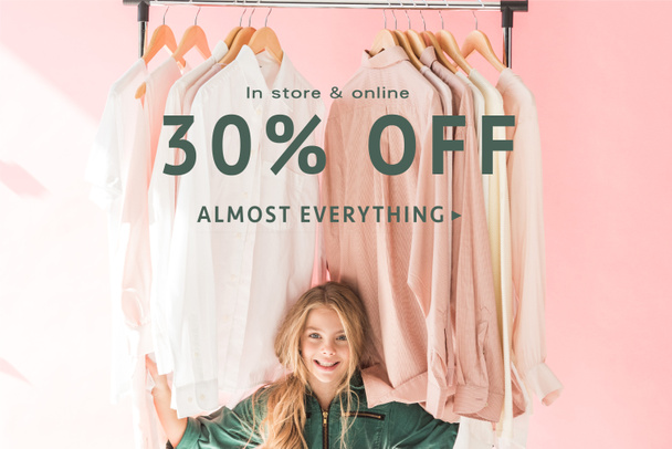 cheerful charming kid in trendy overalls sitting under clothes on hangers, store sale banner concept - Fotó, kép