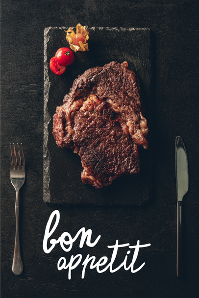 top view of cooked steak and cherry tomato on black wooden board, knife and fork on table in kitchen, bon appetit lettering - Photo, Image