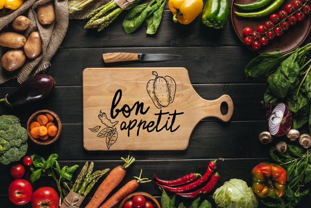 Widok z góry z deska do krojenia z nożem i organicznych warzyw świeżych wokół na drewnianym stole, bon appetit napis - Zdjęcie, obraz