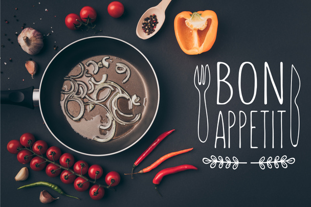top view of frying pan with onion and oil among vegetables on gray table, bon appetit lettering - 写真・画像