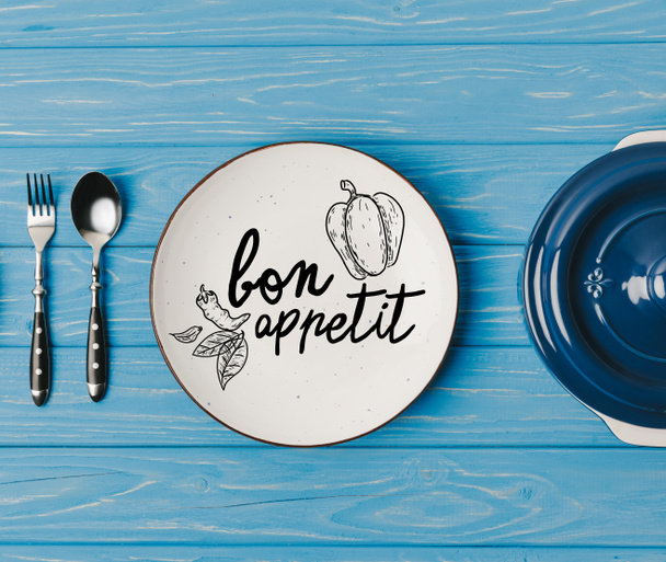 vista superior de tenedor, cuchara y platos en la mesa azul, letras bon appetit
 - Foto, imagen