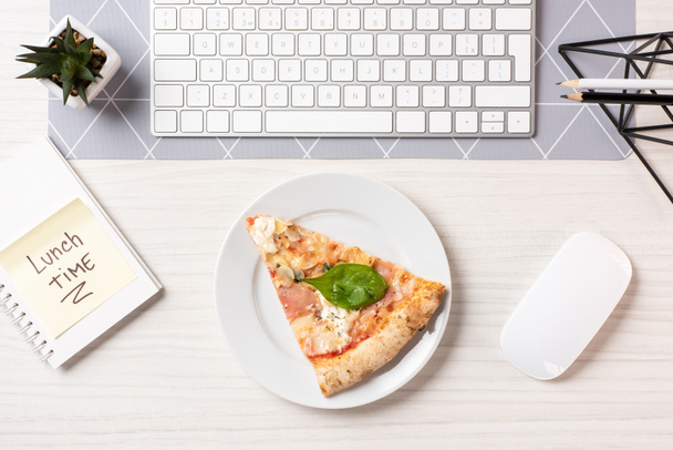 το Top view πίτσα στο πιάτο, σημείωμα με το μεσημεριανό γεύμα χρόνο επιγραφή, υπολογιστή ποντίκι και το πληκτρολόγιο στο χώρο εργασίας - Φωτογραφία, εικόνα