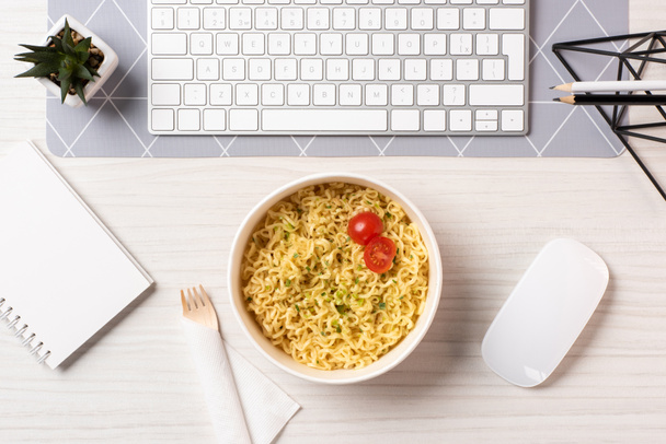 top view of bowl with noodles, fork, computer mouse and keyboard at workplace - Фото, изображение