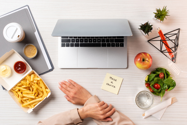 cropped shot of businesswoman, laptop, note with inscription make a choice and healthy food with junk food at workplace  - Fotografie, Obrázek
