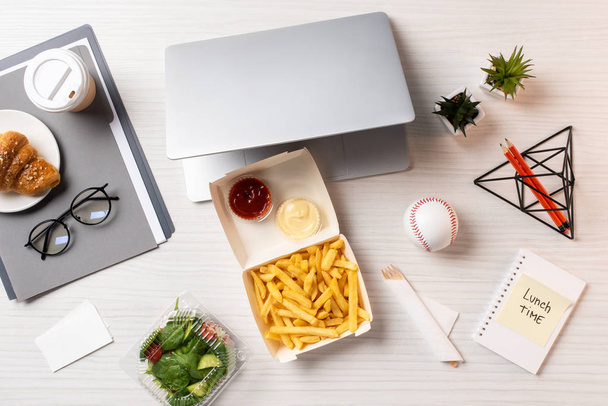 Blick von oben auf Pommes frites, Laptop, Gemüsesalat und Bürobedarf am Arbeitsplatz - Foto, Bild