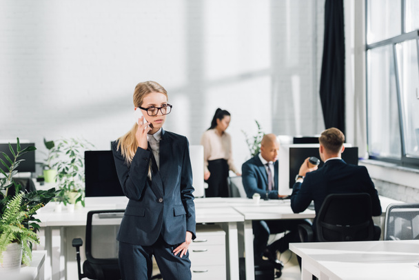 sérieux jeune femme d'affaires parler par smartphone tandis que des collègues travaillant dans un bureau ouvert
 - Photo, image