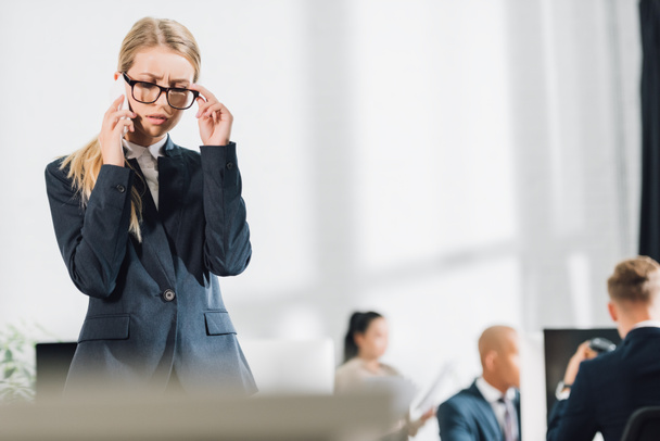 joven empresaria enfocada hablando por teléfono inteligente y ajustando gafas en la oficina
 - Foto, imagen