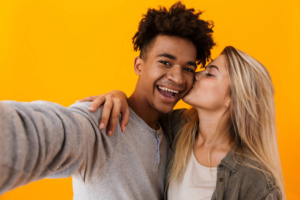 Image of happy cute young loving couple posing isolated over yellow background hugging take a selfie by camera kissing. - Foto, Imagem